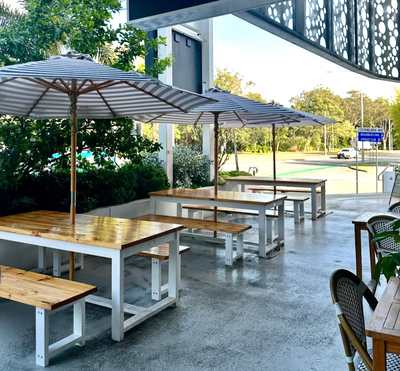 Outdoor seating area with wooden tables and benches under large umbrellas, surrounded by greenery.